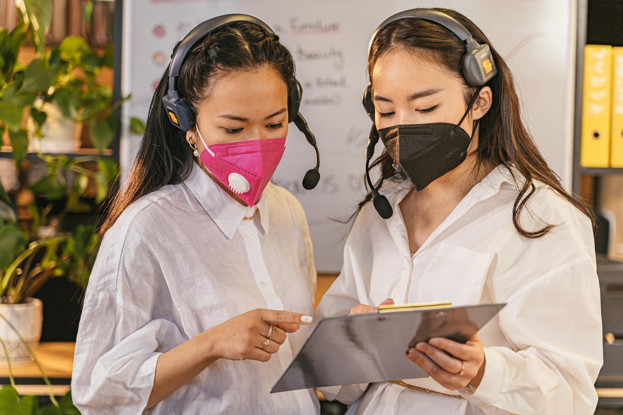 Two women in face masks and headsets discussing work on a clipboard in an office setting.